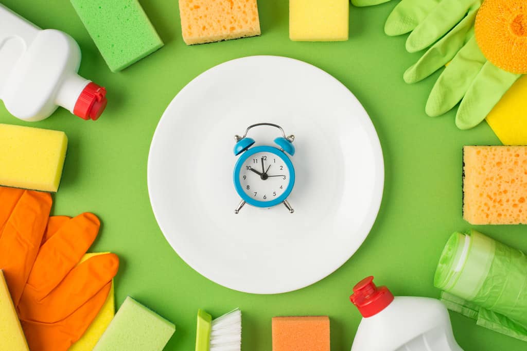 Top view photo of blue alarm clock on white clean plate in the middle detergent bottles rubber gloves rags sponges brush and garbage bags on isolated green background